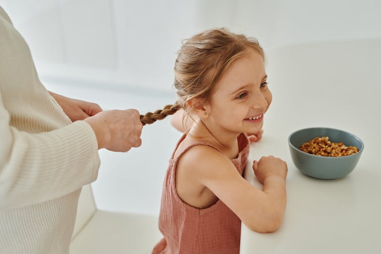 A mother braiding her smiling daughter's hair beside a bowl of cereal in a warm kitchen.