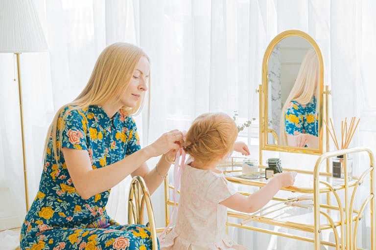 A woman braids her daughter's hair at a vanity mirror in a bright room.