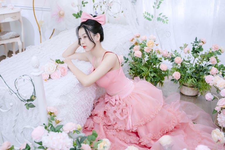 A woman in a pink gown with a hair bow, surrounded by flowers, poses gracefully by a bed indoors.