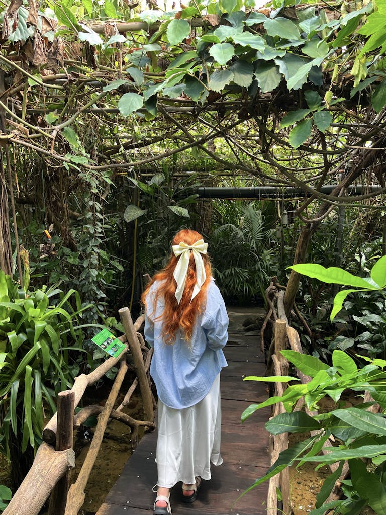 A woman with long red hair and a hair bow walks in a jungle path.