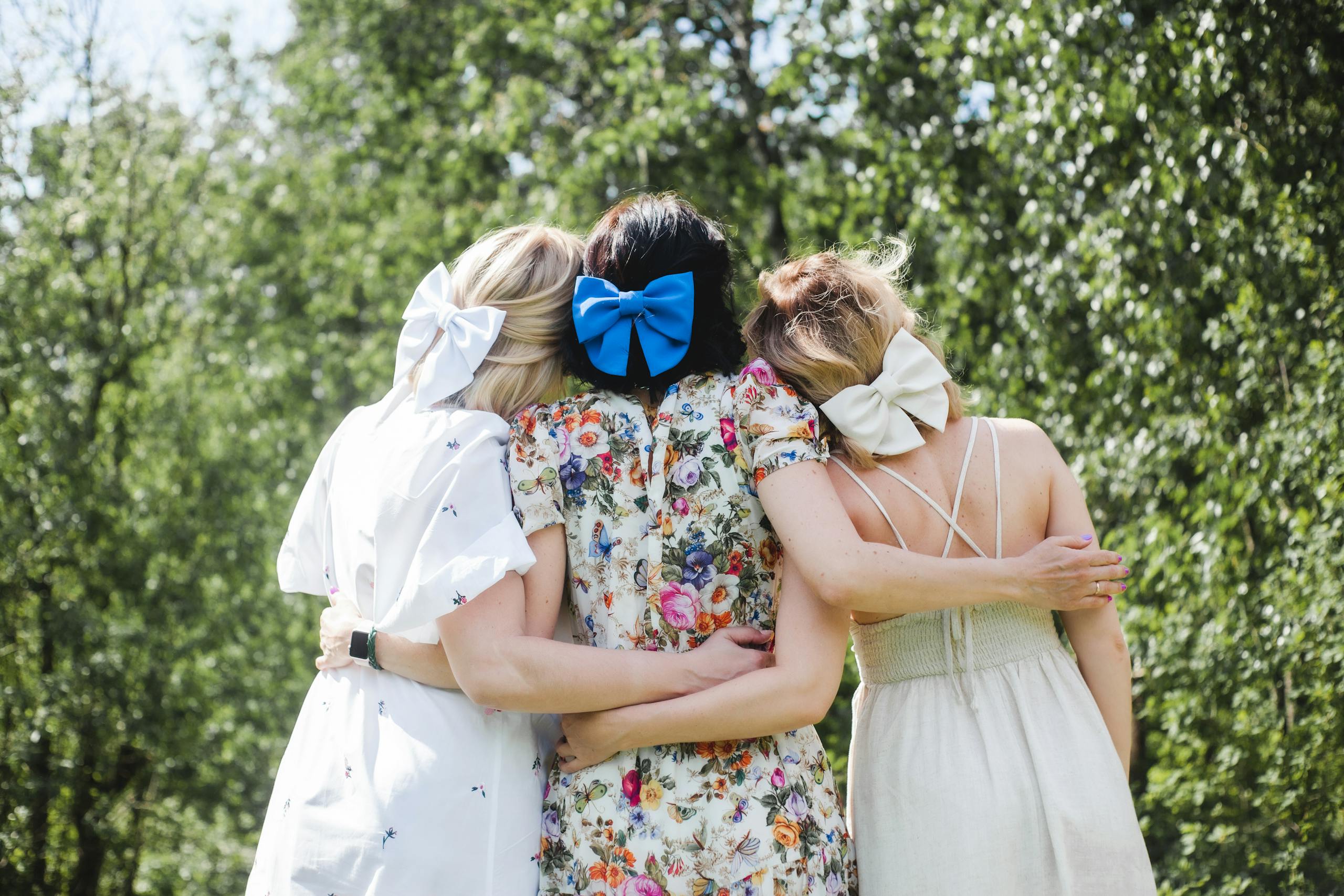 Three women with bows in their hair embrace outdoors, showcasing friendship and summer vibes.