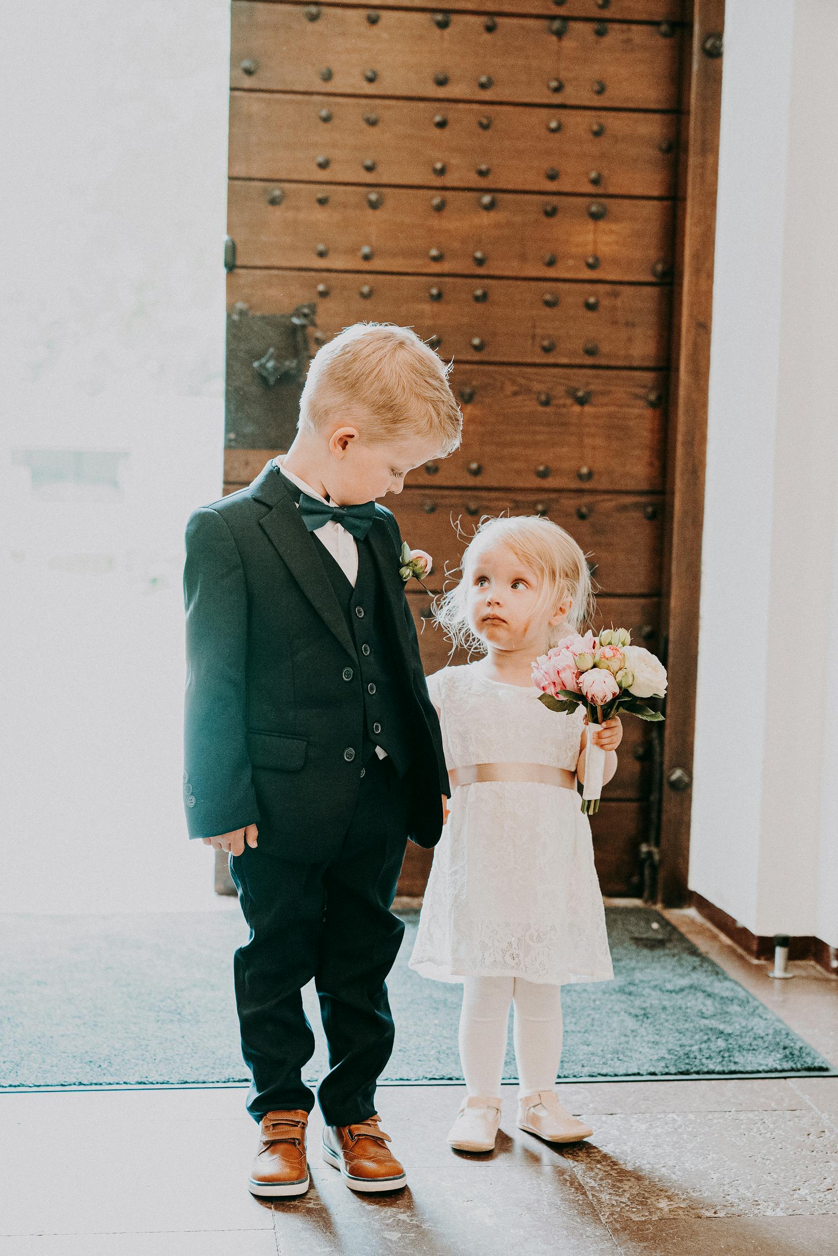 Full body of little girl in white bridal gown holding wedding bouquet and looking at boy in black elegant tuxedo in sunlight