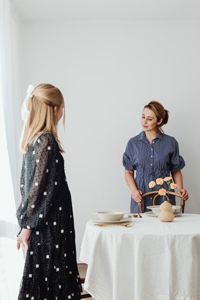 A mother and daughter setting up a dining table at home, with tableware and flowers.