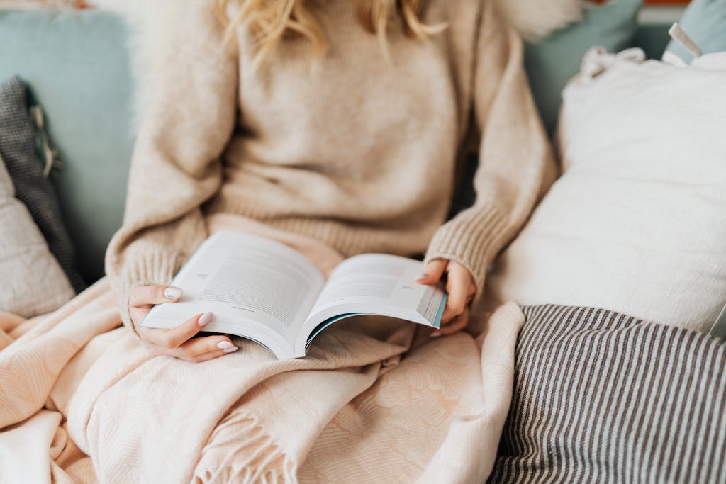 A person with manicured nails enjoys a book while wrapped in a cozy blanket.