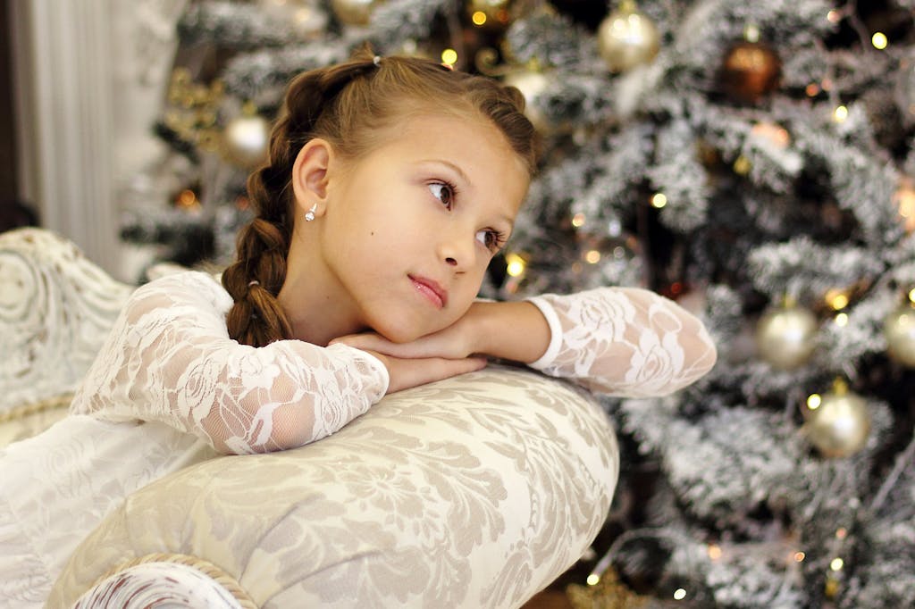 A thoughtful young girl rests on a sofa beside a beautifully decorated Christmas tree indoors.