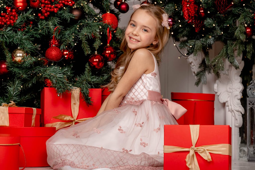 A young girl in a dress poses with gifts under a Christmas tree, creating a festive scene.