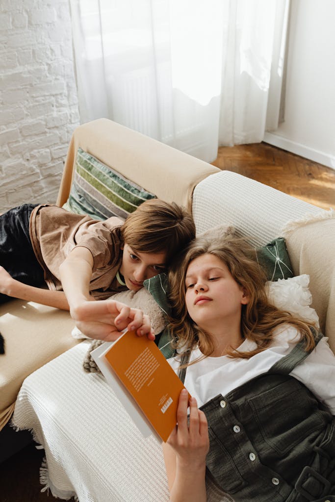 Two teenagers reading together on a sofa indoors, creating a warm and relaxed atmosphere.