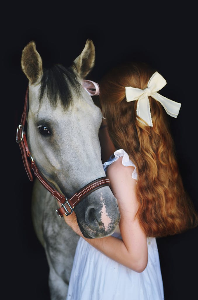 Young girl with red hair and bow embraces a white horse on a black background.