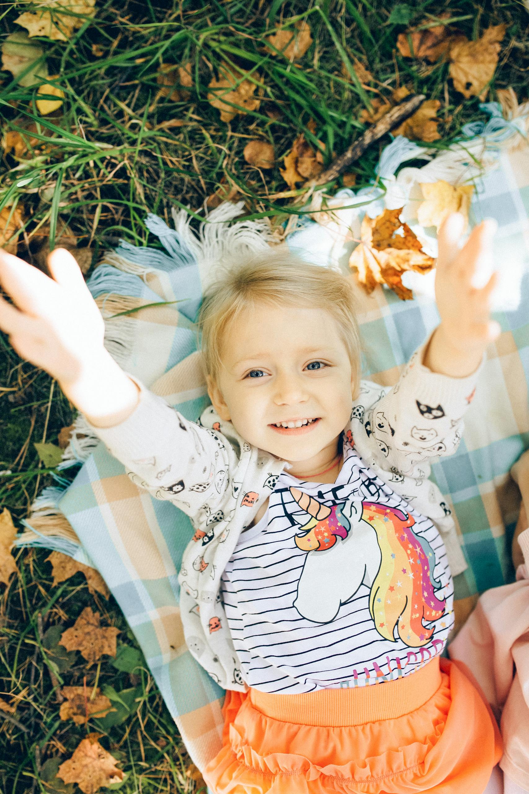 A cheerful young girl with blonde hair lays on a blanket outdoors, embracing the autumn vibes.