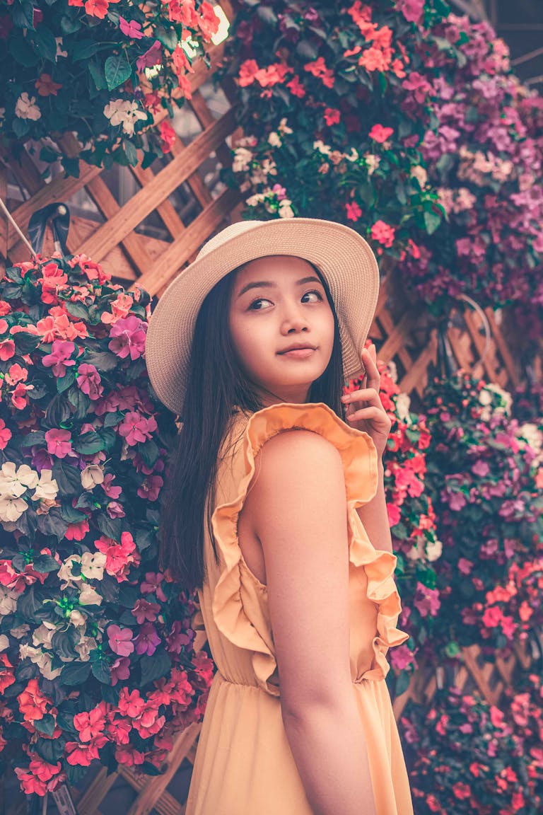 Stylish Asian teenager wearing a yellow dress and hat posing in front of a vibrant floral background.